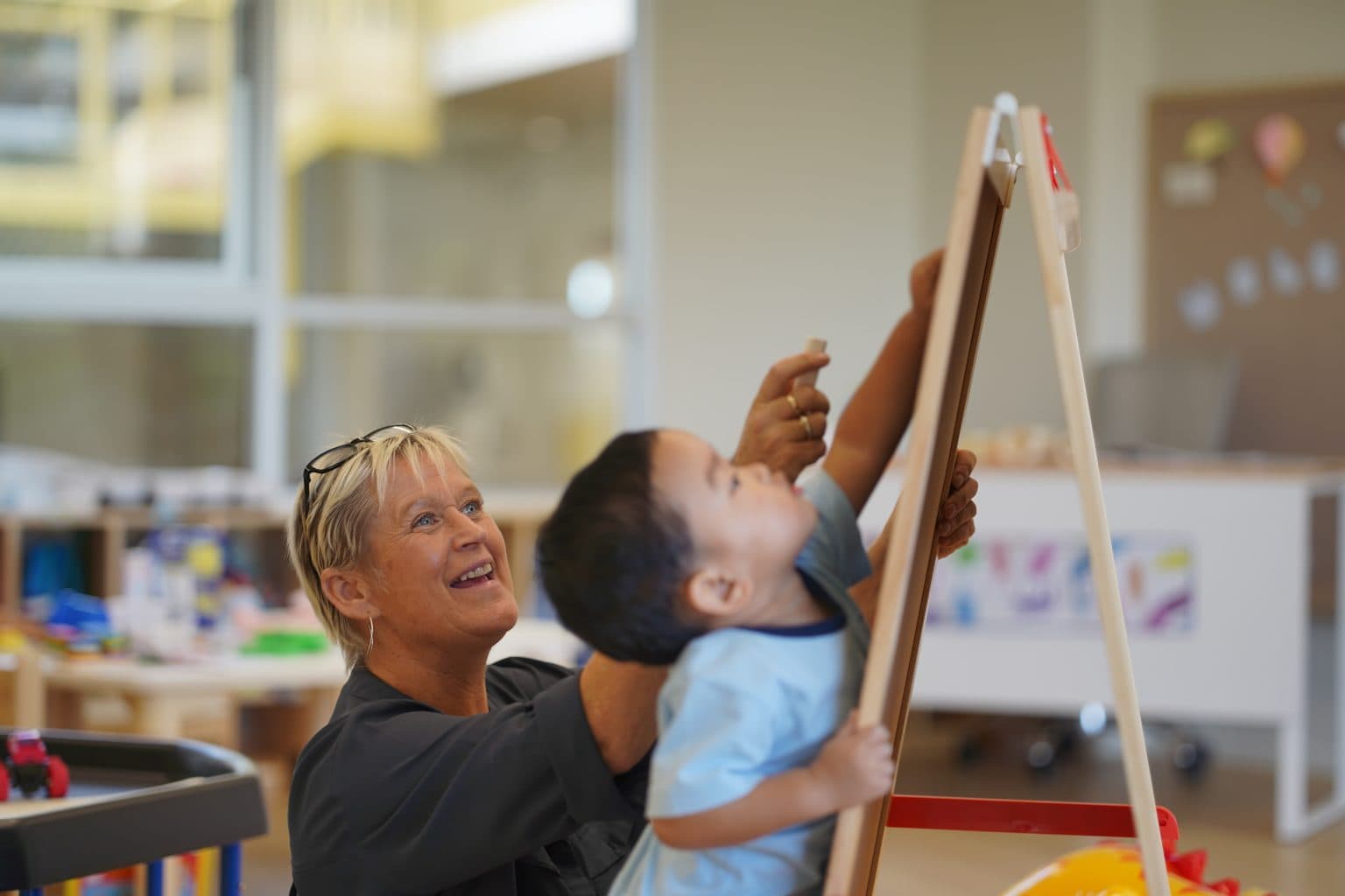 A Toddler reaching up to the easel while his teacher smiles